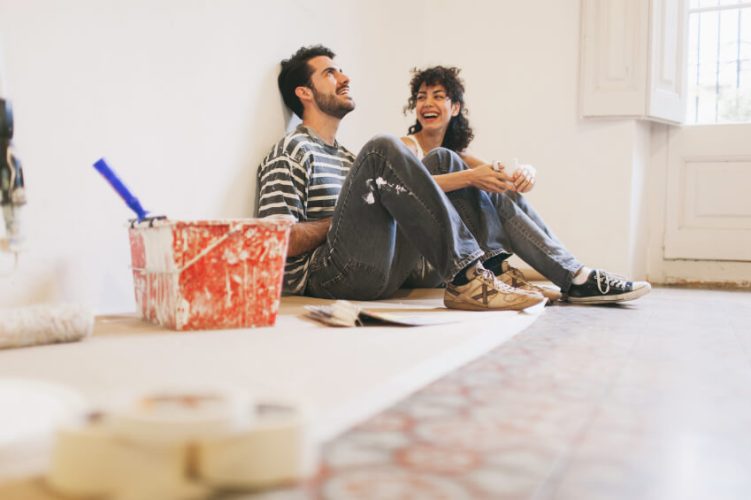 Young couple laughing sitting on floor of new house next to a paint bucket.