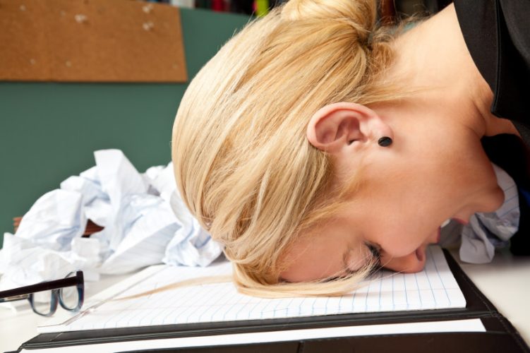 Young upset female employee resting head on desk next to bunch of crumpled papers and her glasses