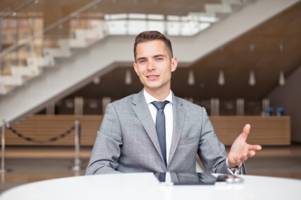 Young businessman extending left hand over table talking about high deductible. Office staircase and main counter behind him.