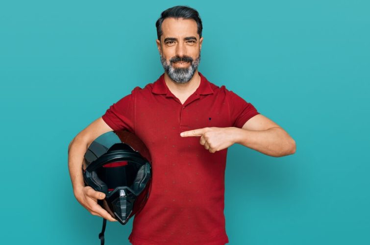 Man in red shirt with blue background stands and points to his motorcycle helmet - cheap motorcycle insurance.