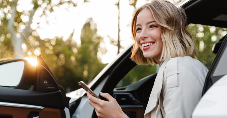 Woman smiling and watching her phone besides her car thinking about getting Auto refinancing