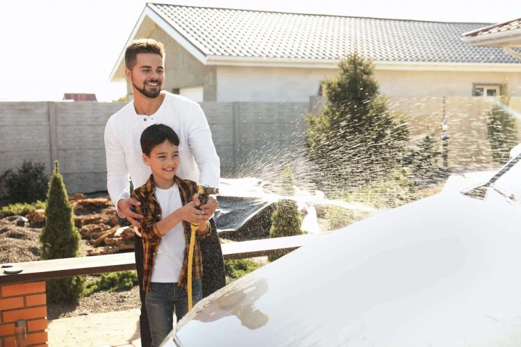 Smiling young father with his kid washing a car with a water hose.