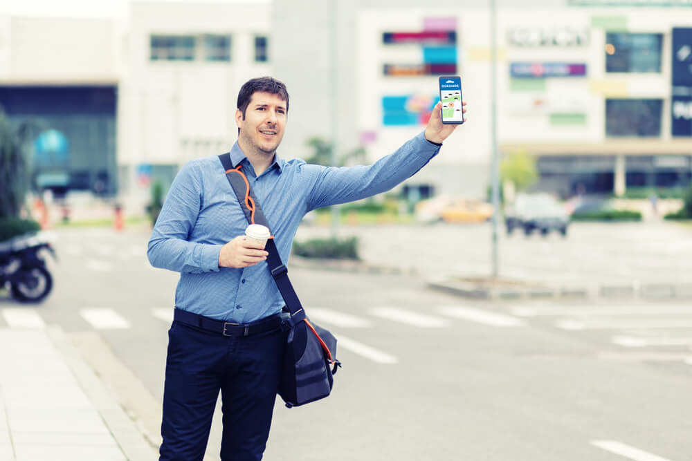 Man standing on side of road holding his phone out to alert his Lyft driver - cheap rideshare insurance.