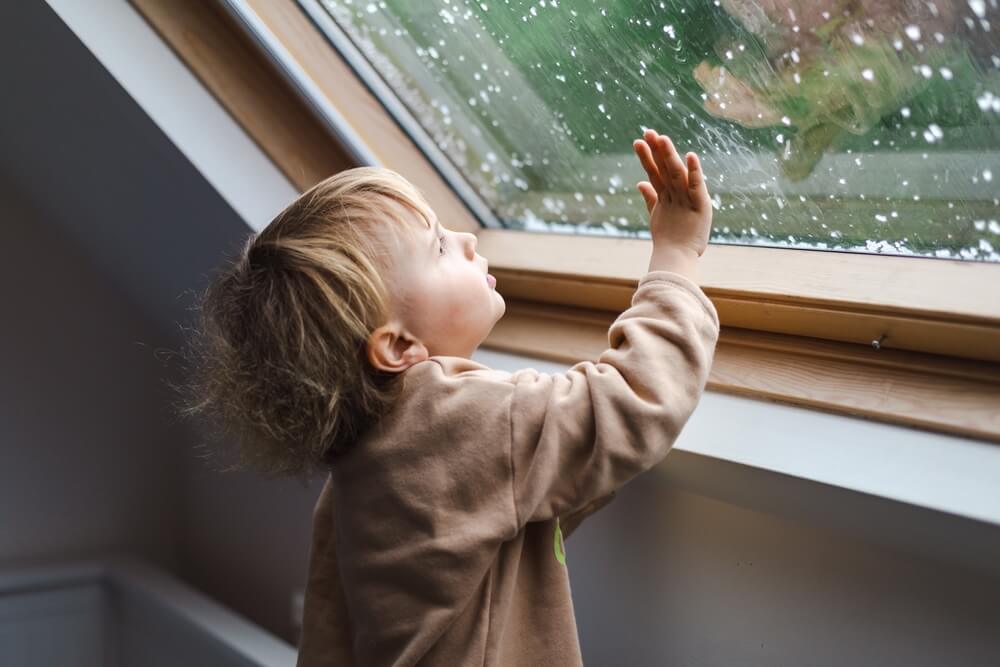 Little preschool boy staying home in bad weather and looking with interest through window on raindrops and hail, indoors.