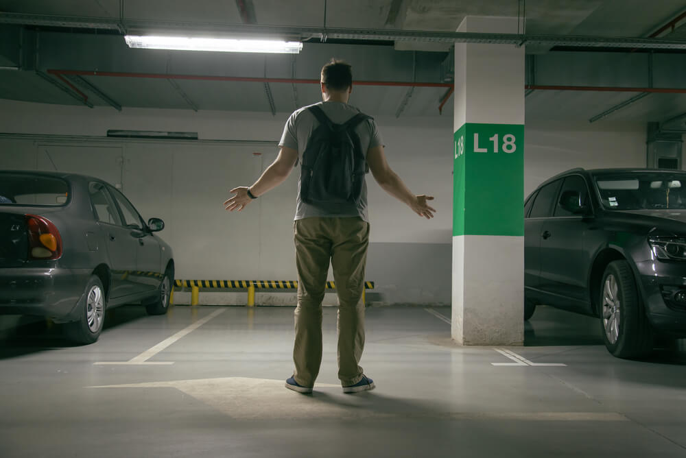 Puzzled young man with backpack, back to camera, looks at his empty parking space where his car used to be.