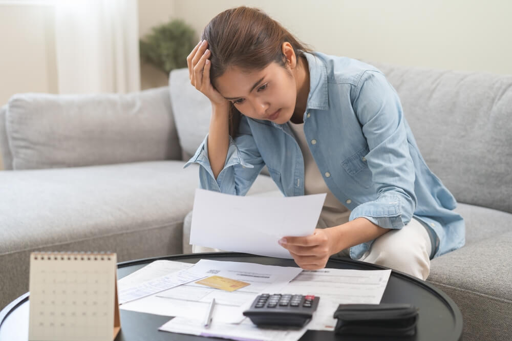 Rising car insurance costs, stressed Asian young woman have a headache while sitting on sofa calculate expenses, income - cheap car insurance in California.