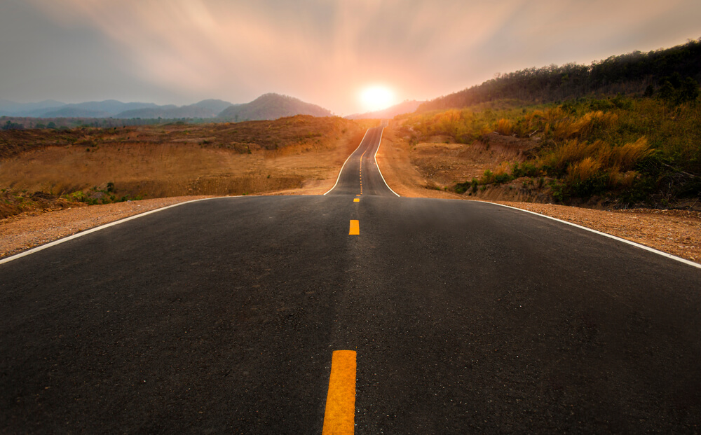 A two-lane highway leads to the horizon with the sun setting - cheap car insurance.